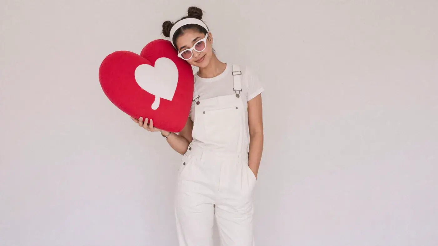 A young woman in white dungarees hugs a red heart cushion against a pale wall. Sample image created by Quiamo, a brand, marketing and media agency from India.