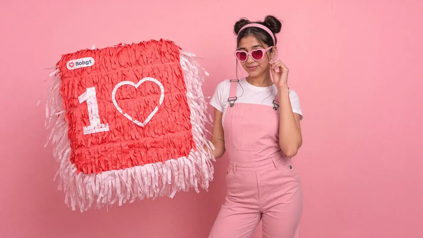 A young woman in pink dungarees and heart sunglasses holds a giant like-counter pillow against pink. Sample image created by Quiamo, a brand, marketing and media agency from India.