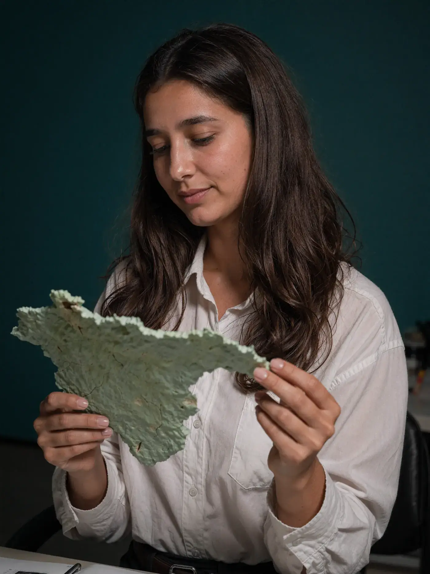 A woman in a white shirt studies a sheet of textured green handmade paper against deep teal. Sample image created by Quiamo, a brand, marketing and media agency from India.