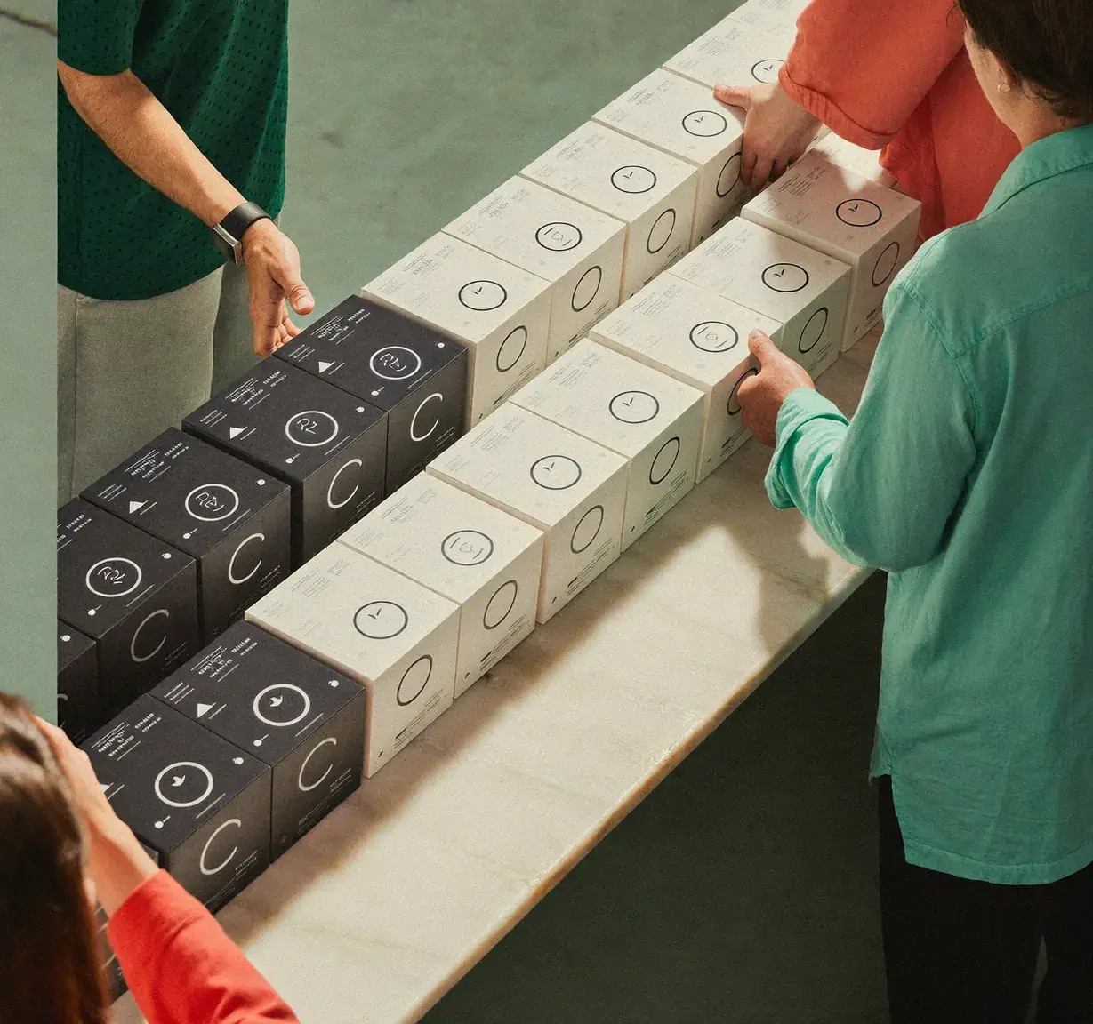 A team packs rows of branded white and black boxes on a long table, seen from above. Sample image created by Quiamo, a brand, marketing and media agency from India.