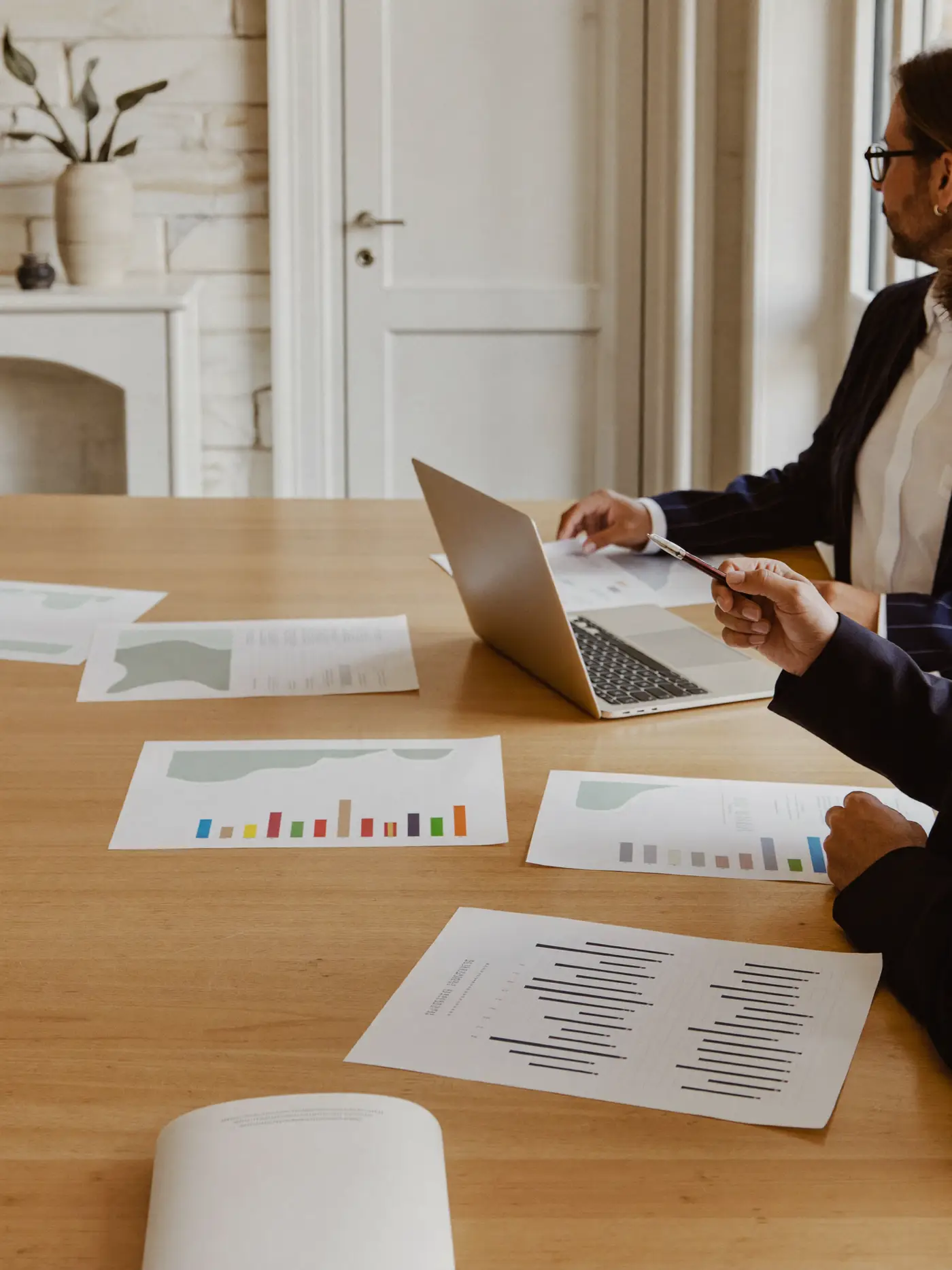 Two professionals reviewing printed charts and a laptop in a bright advisory meeting room.