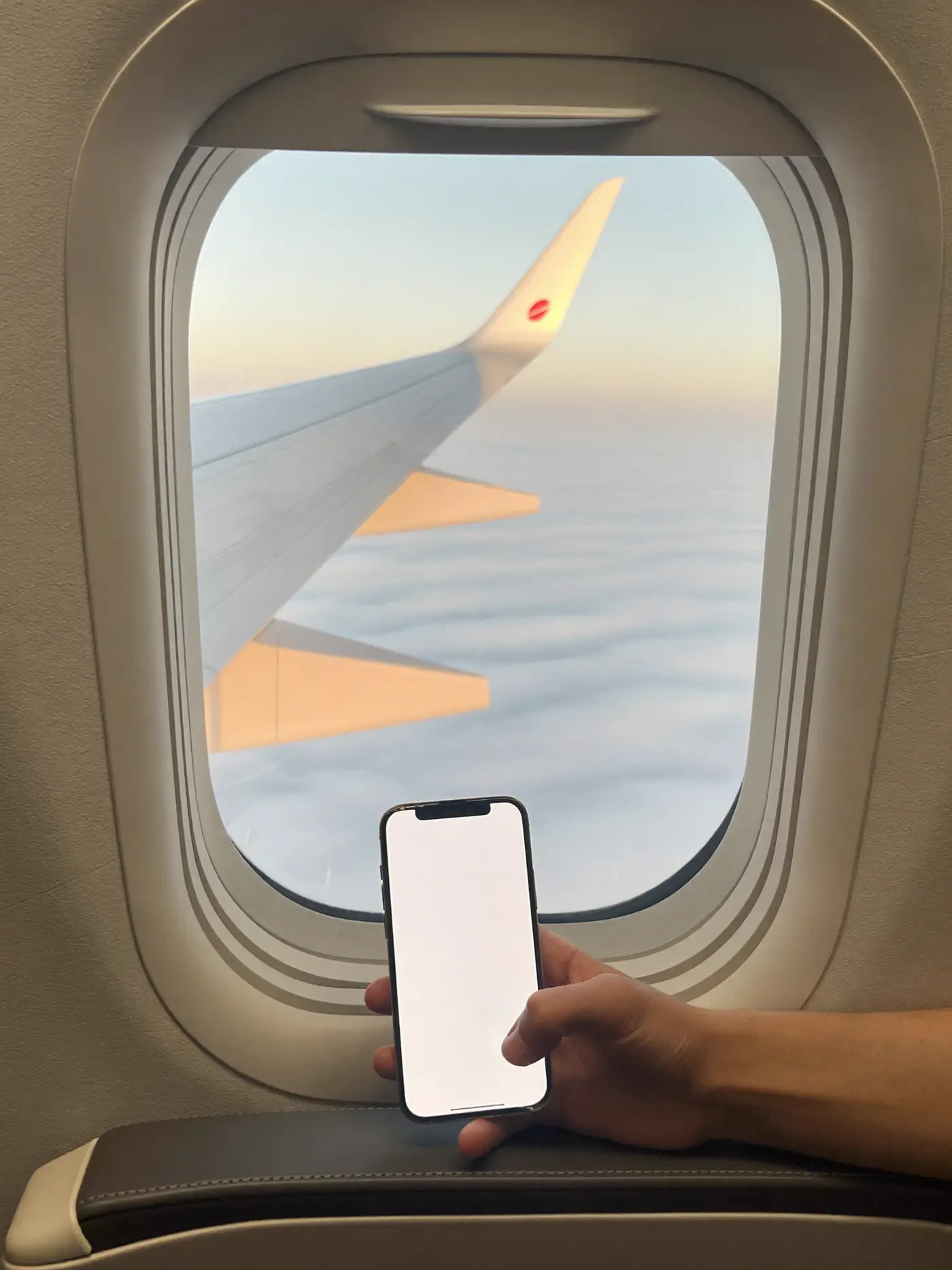 A traveller's hand holding a phone beside an aircraft window showing the wing above the clouds.