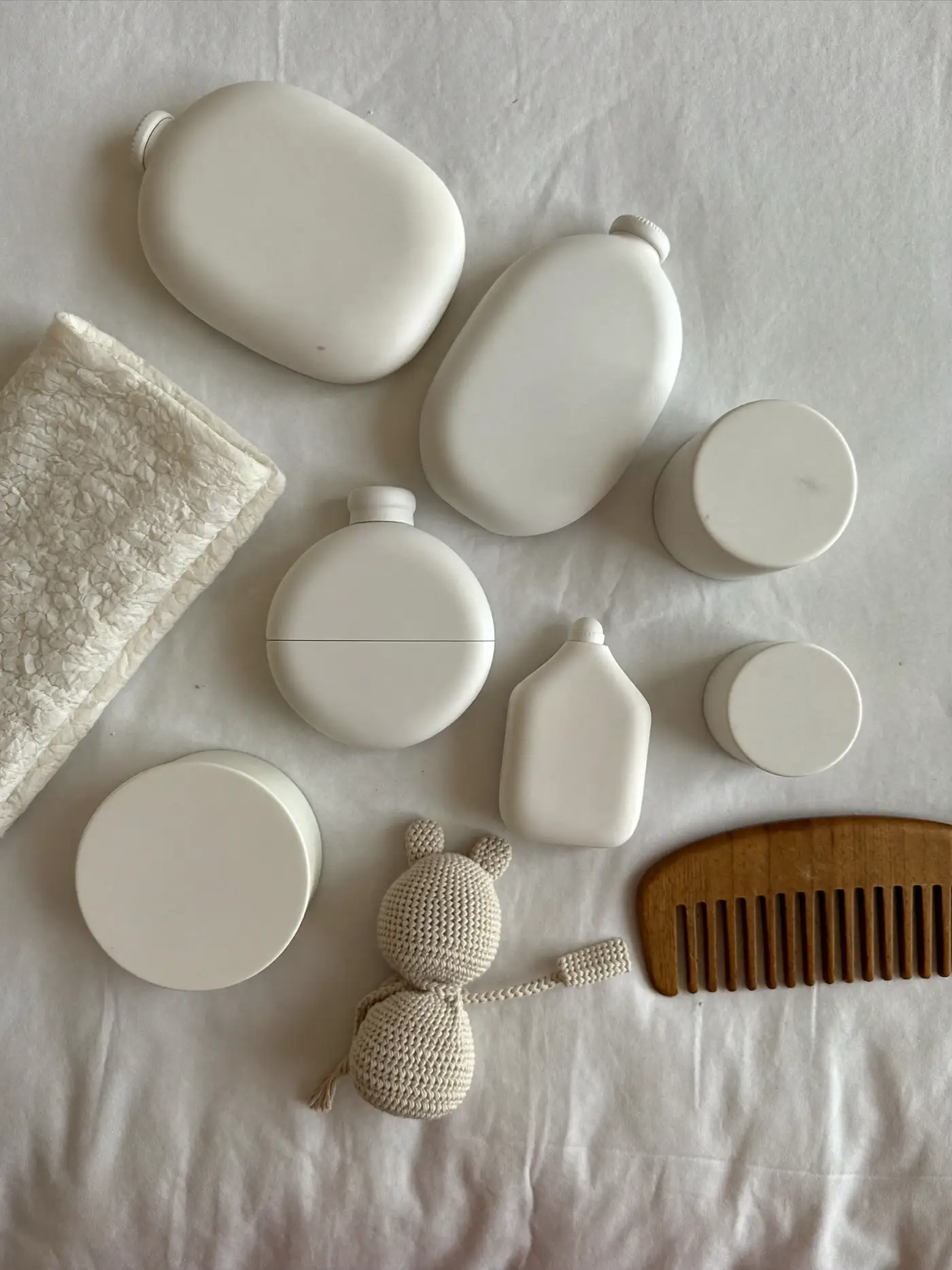 A soft flat lay of plain white baby care bottles and jars with a muslin cloth, a knitted toy and a wooden comb.