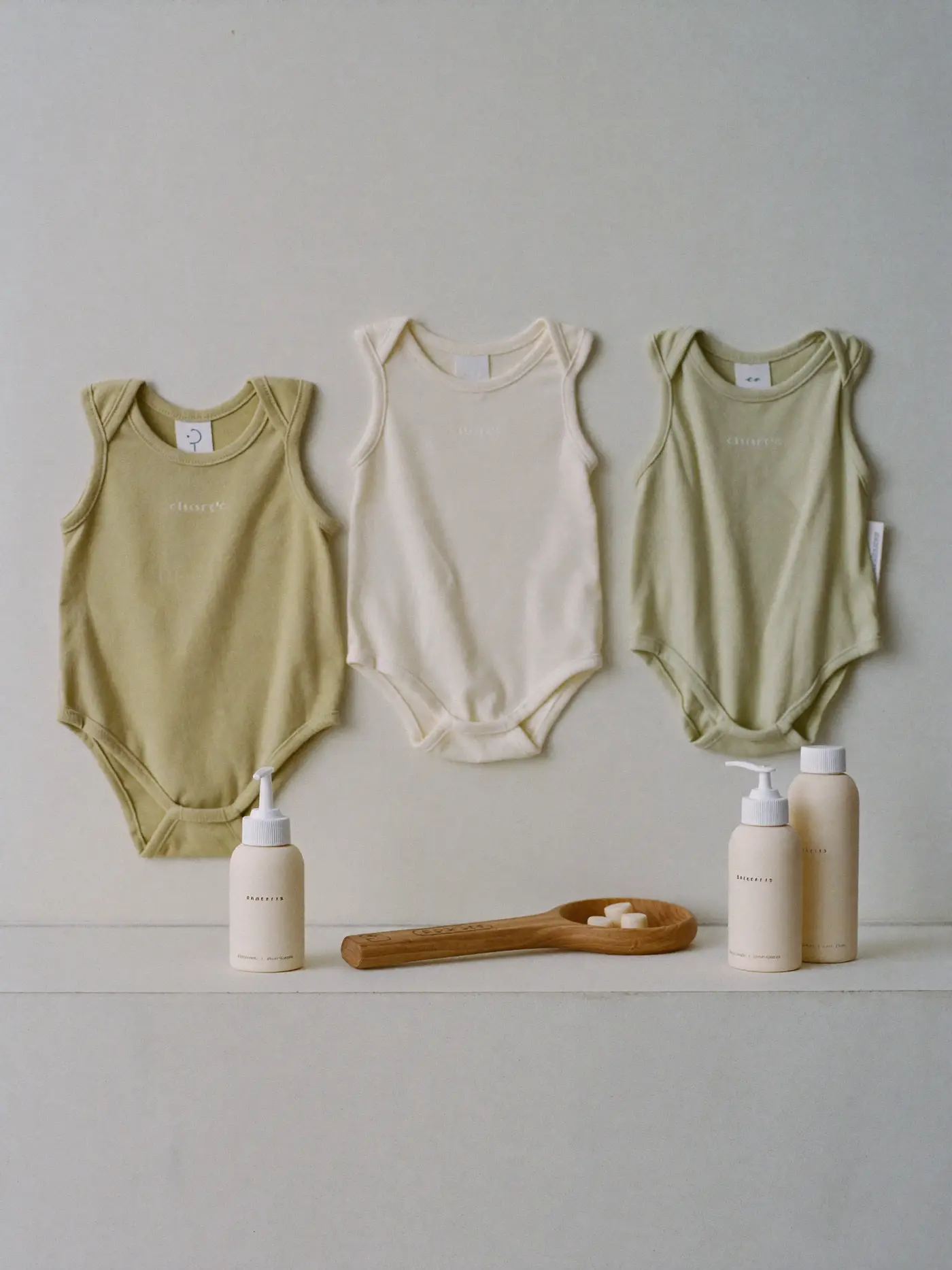 Baby onesies in sage and cream with a wooden rattle and bottles, set up for catalogue photography.
