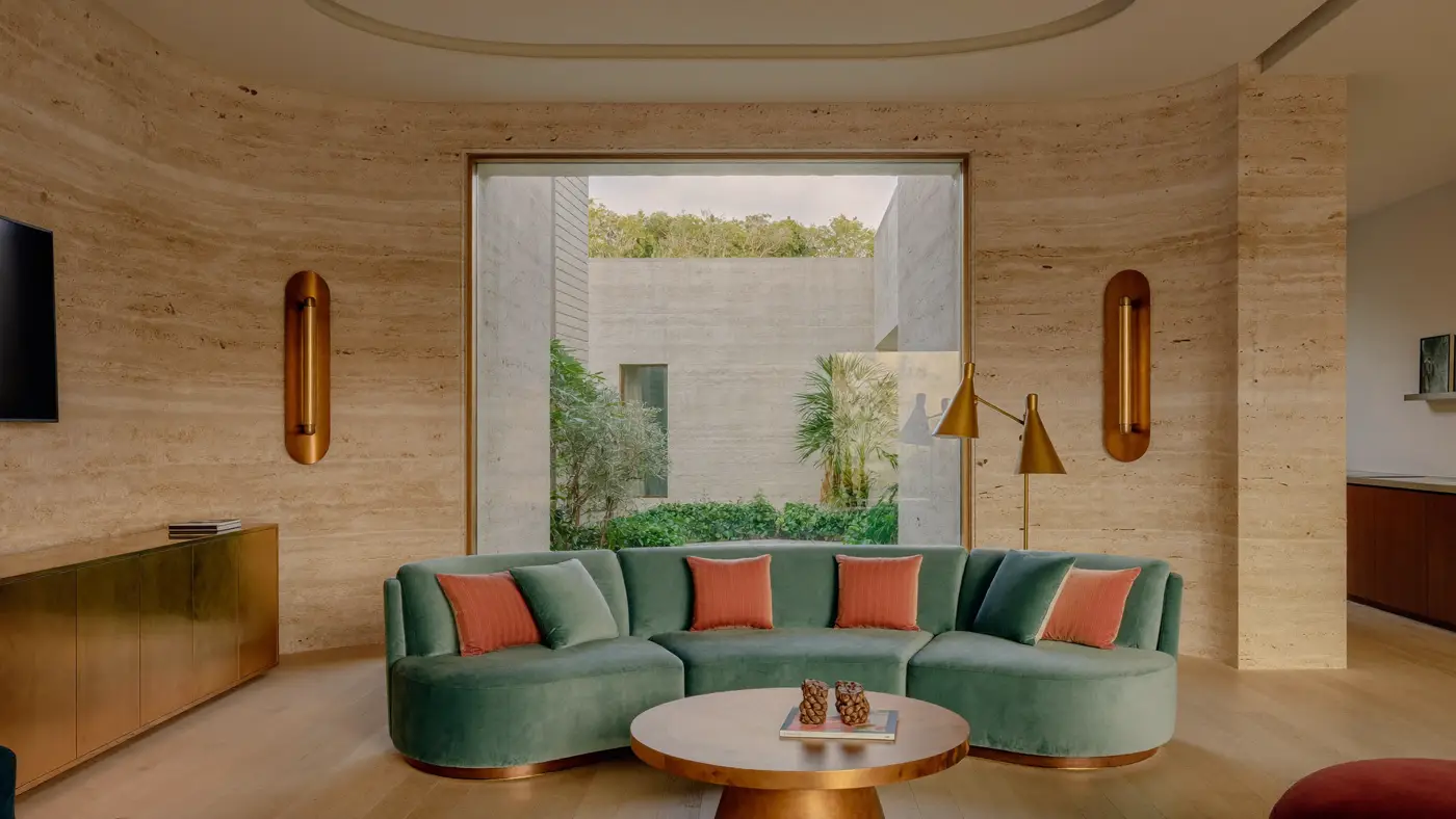 A boutique hotel lobby with travertine walls, a curved teal sofa with coral cushions and a garden courtyard view.