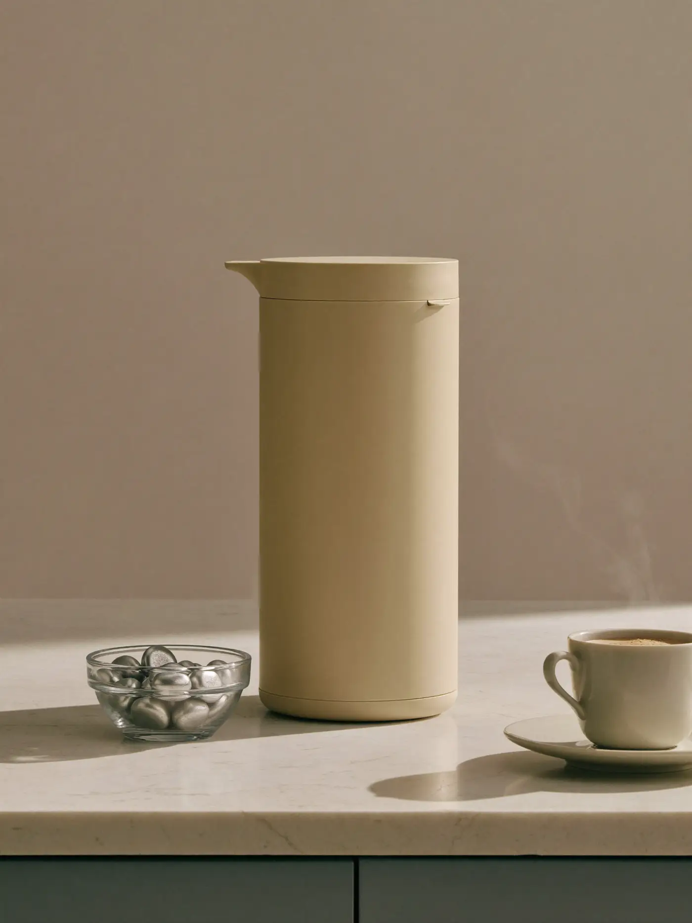 A cream capsule coffee machine on a limestone counter with a bowl of silver capsules and a cup of coffee.