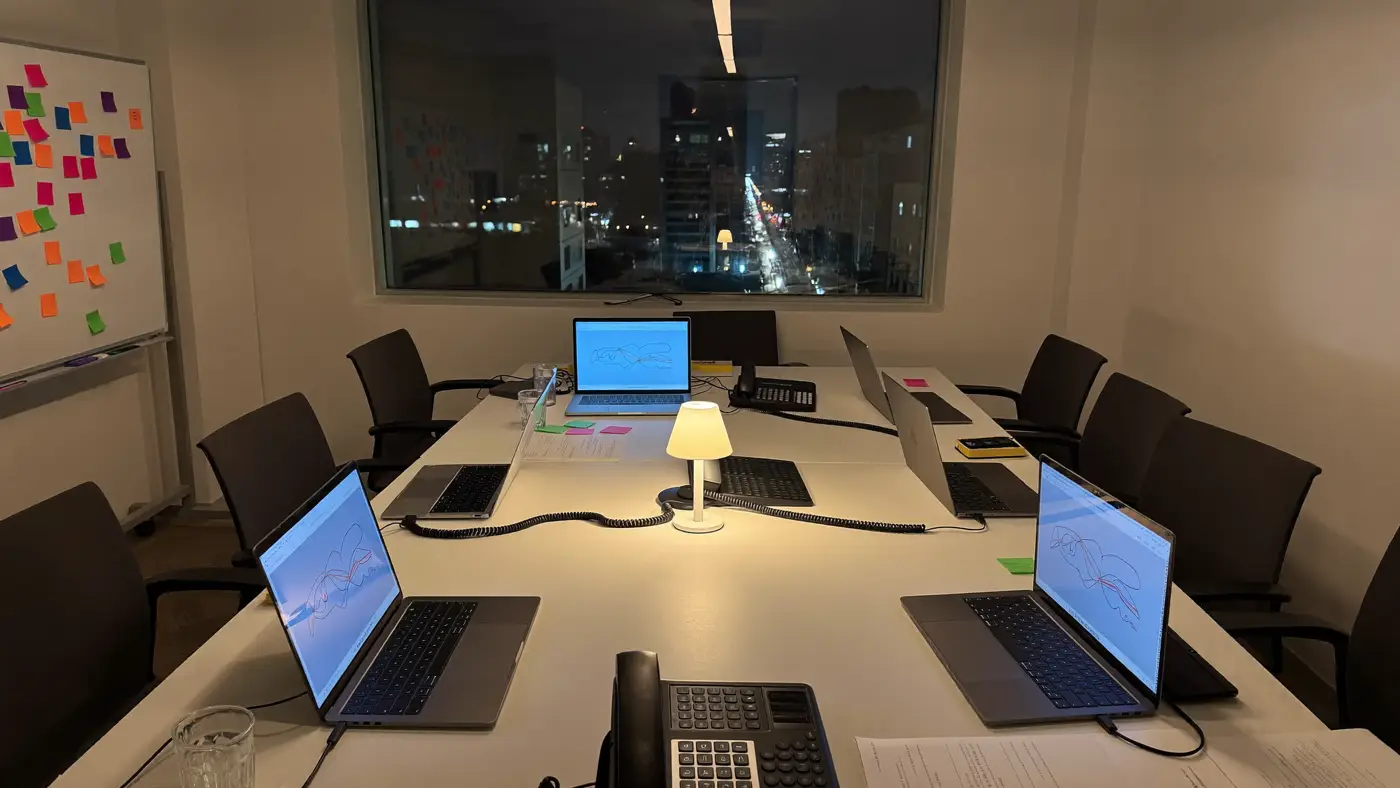 A communications room at night with laptops on a long table, a wall of coloured notes and city lights outside.