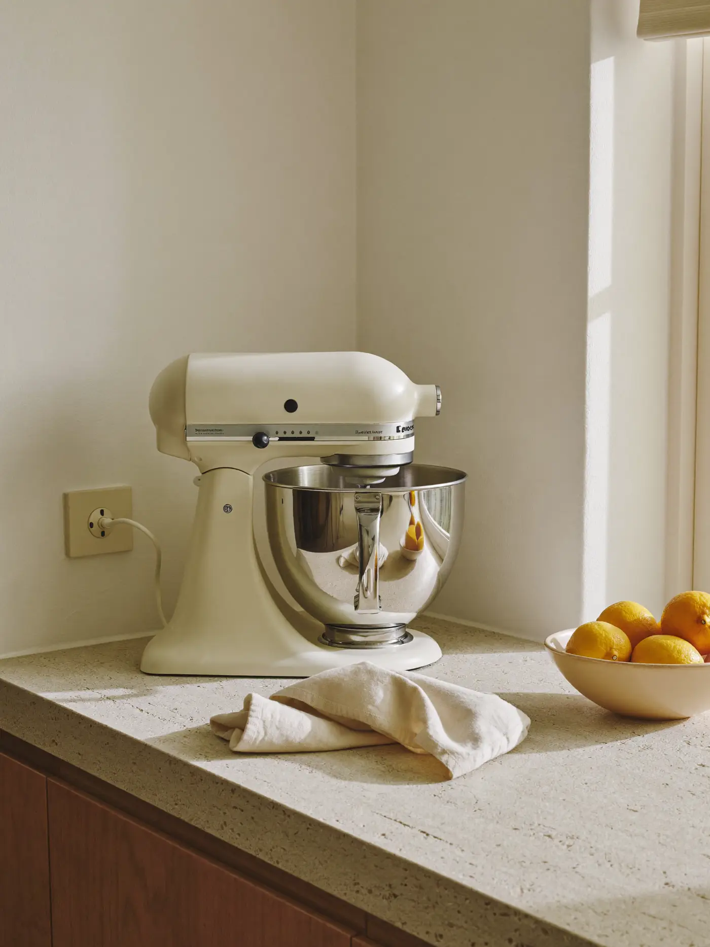A cream stand mixer with a steel bowl on a limestone kitchen counter beside a bowl of lemons in morning sun.