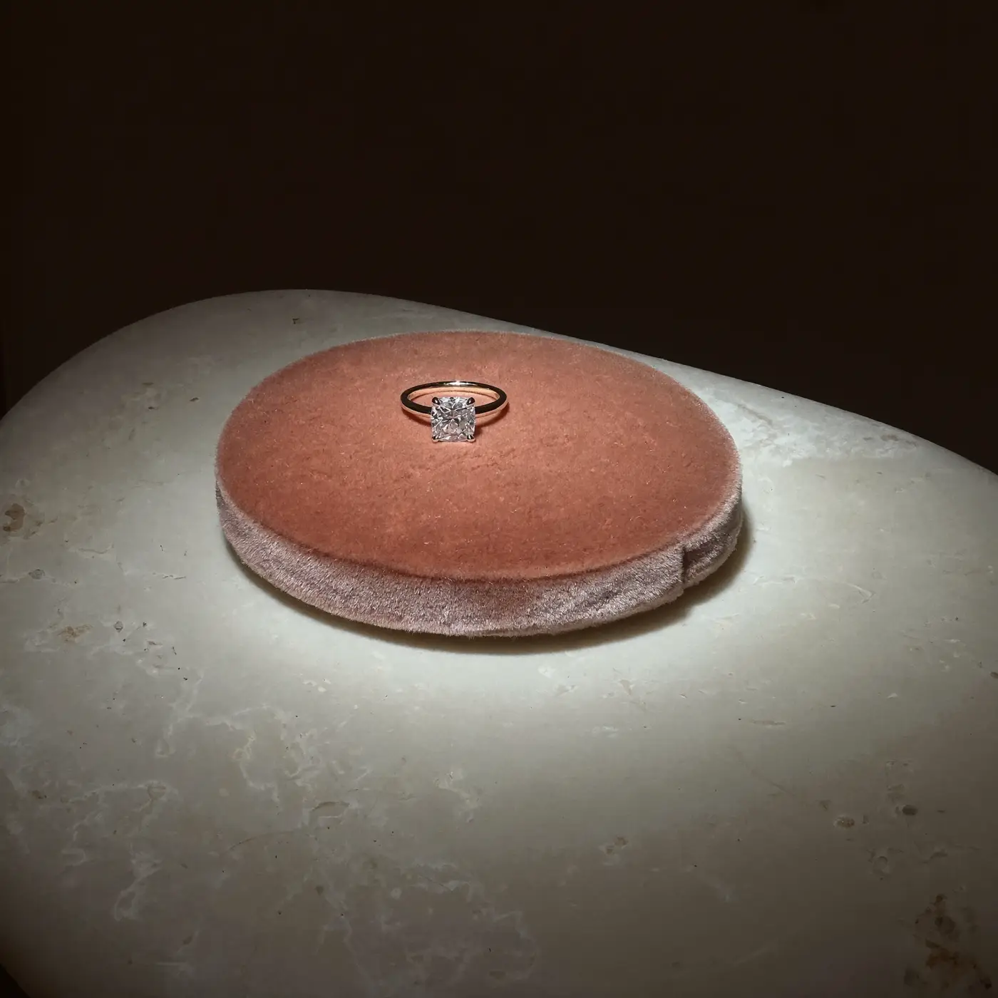 A diamond solitaire ring on a rose velvet plinth under a single spotlight.