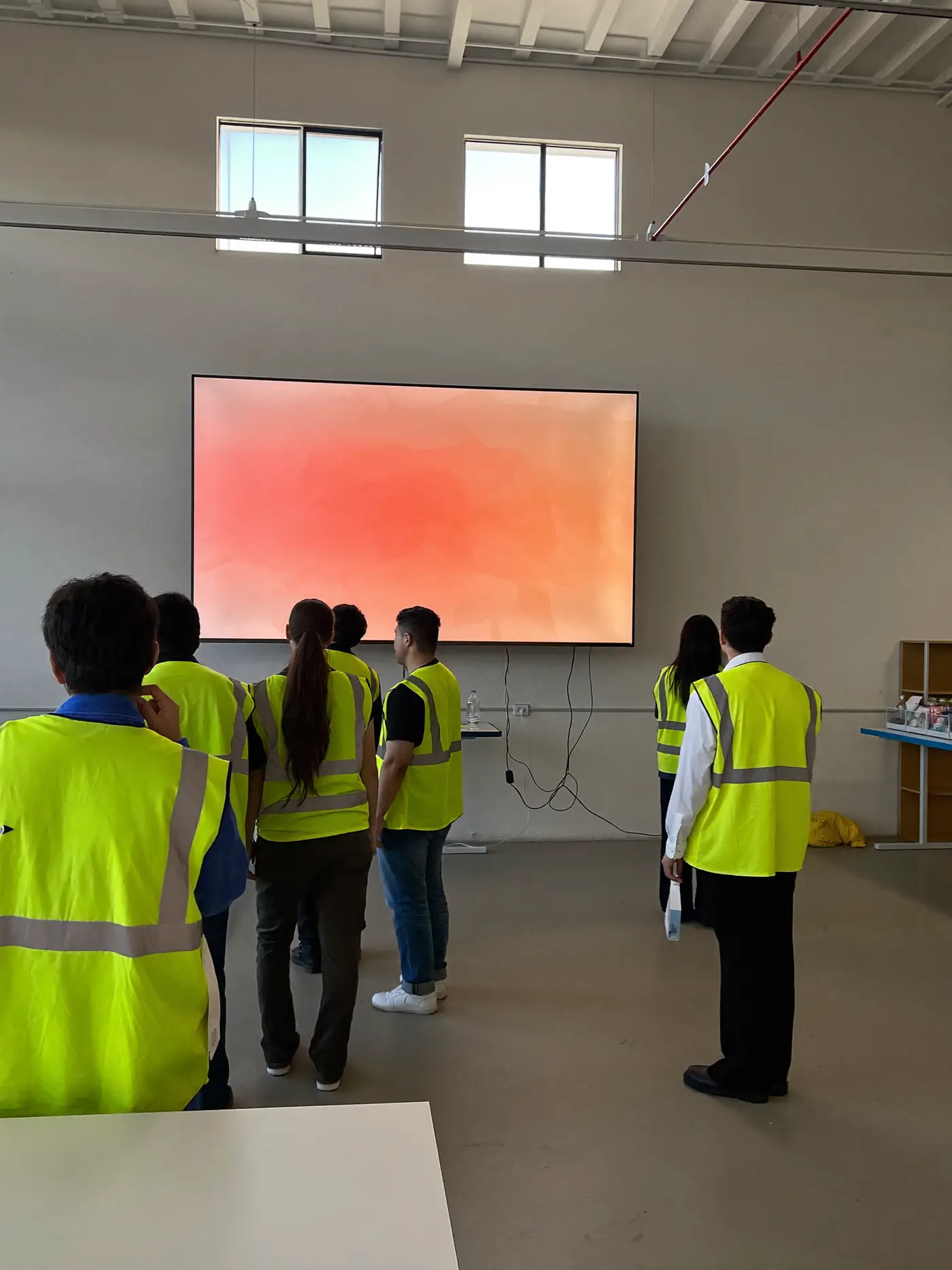 Factory team members in hi-vis vests watching a large screen together in a bright canteen.