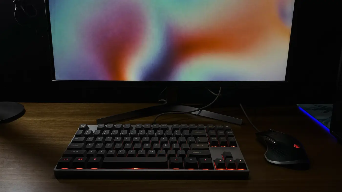A low-profile mechanical keyboard and gaming mouse on a walnut desk at night below a glowing monitor.