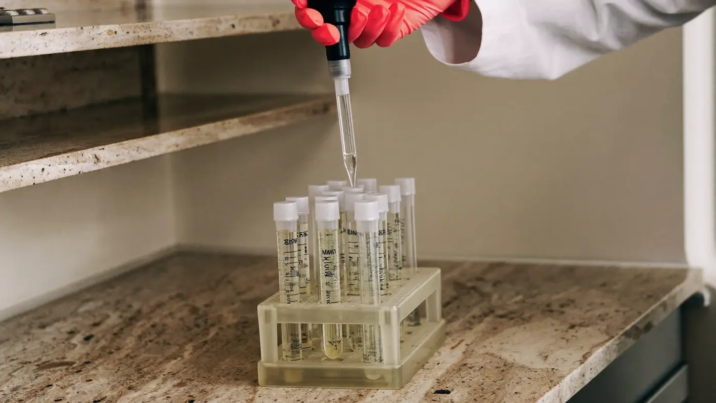 A gloved hand using a glass pipette over a rack of test tubes on a travertine laboratory bench.