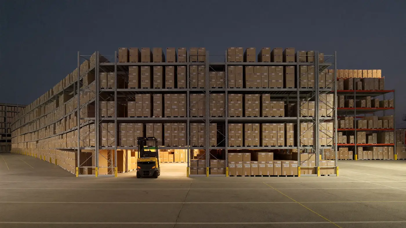 A modern logistics warehouse at dusk with tall shelving of plain boxes and a forklift under amber light.