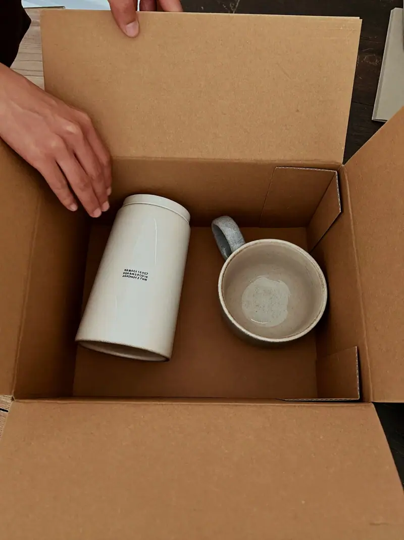 Hands packing a ceramic cup into a kraft box beside a well-used identical cup, illustrating customer lifetime value.