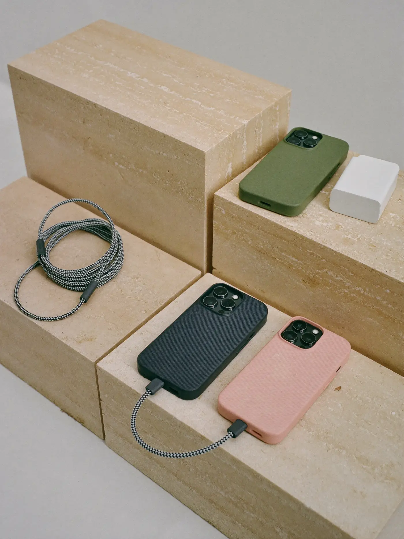 Phone cases in sage, blush and graphite with braided cables and a white charger arranged on stepped limestone plinths.