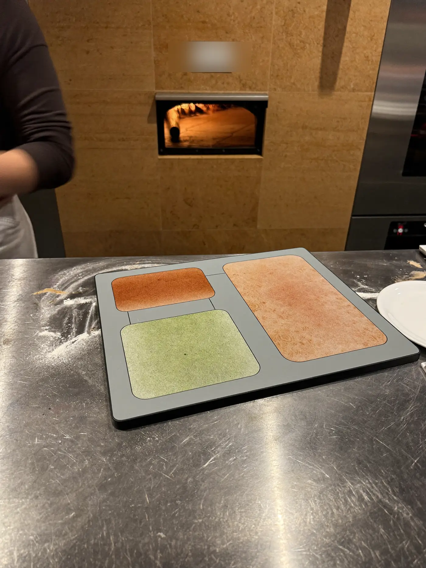 A tablet showing a simple order flow on a steel pizza kitchen counter beside a wood-fired oven.