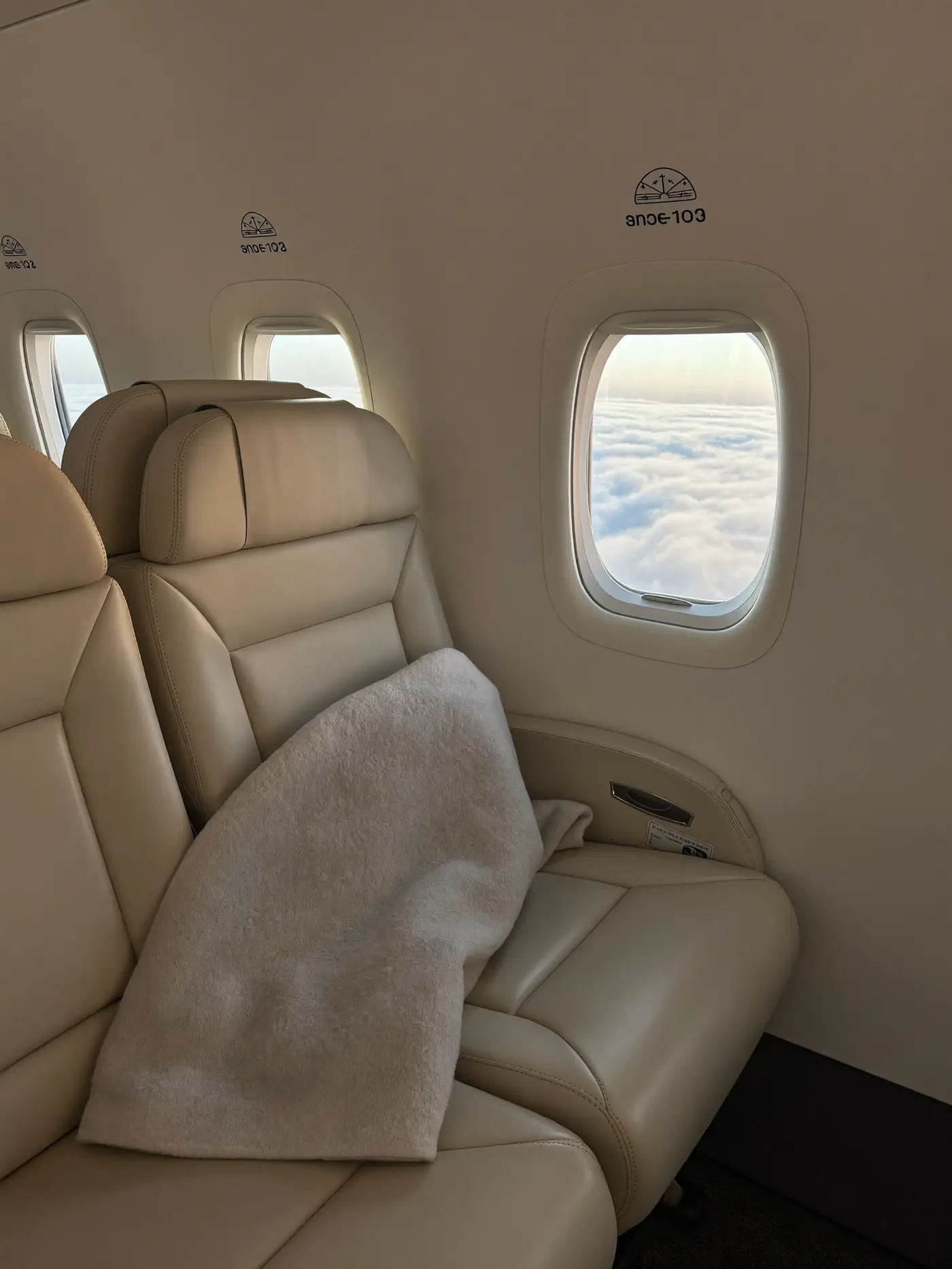 A calm premium aircraft cabin with cream leather seats and clouds at sunrise through the window.