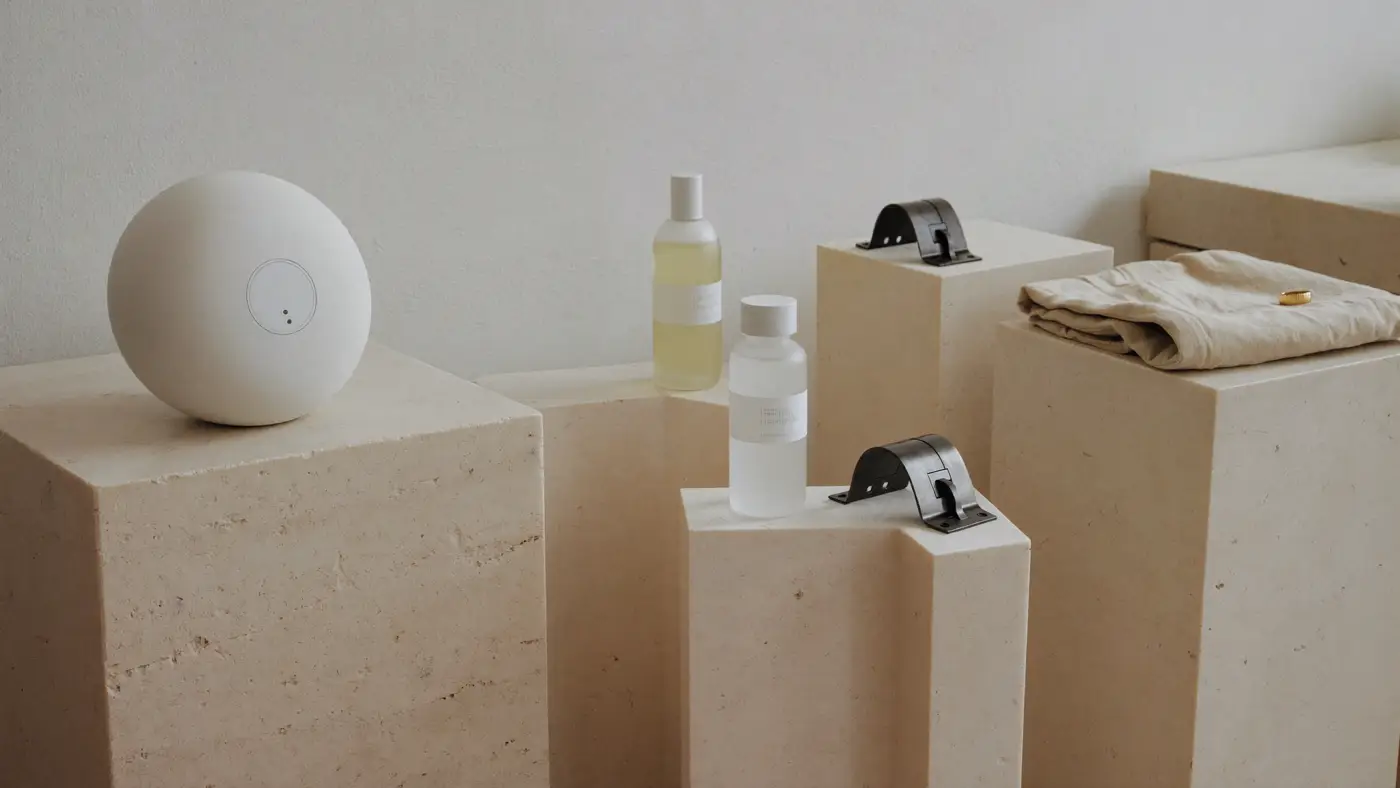 A white speaker, glass bottles, machined steel brackets and a folded linen shirt on limestone plinths.