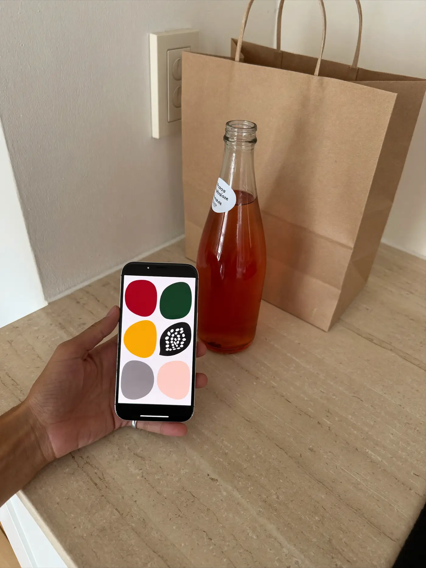 A hand holding a phone with a colourful app grid beside a bottle of pink drink and a paper delivery bag.
