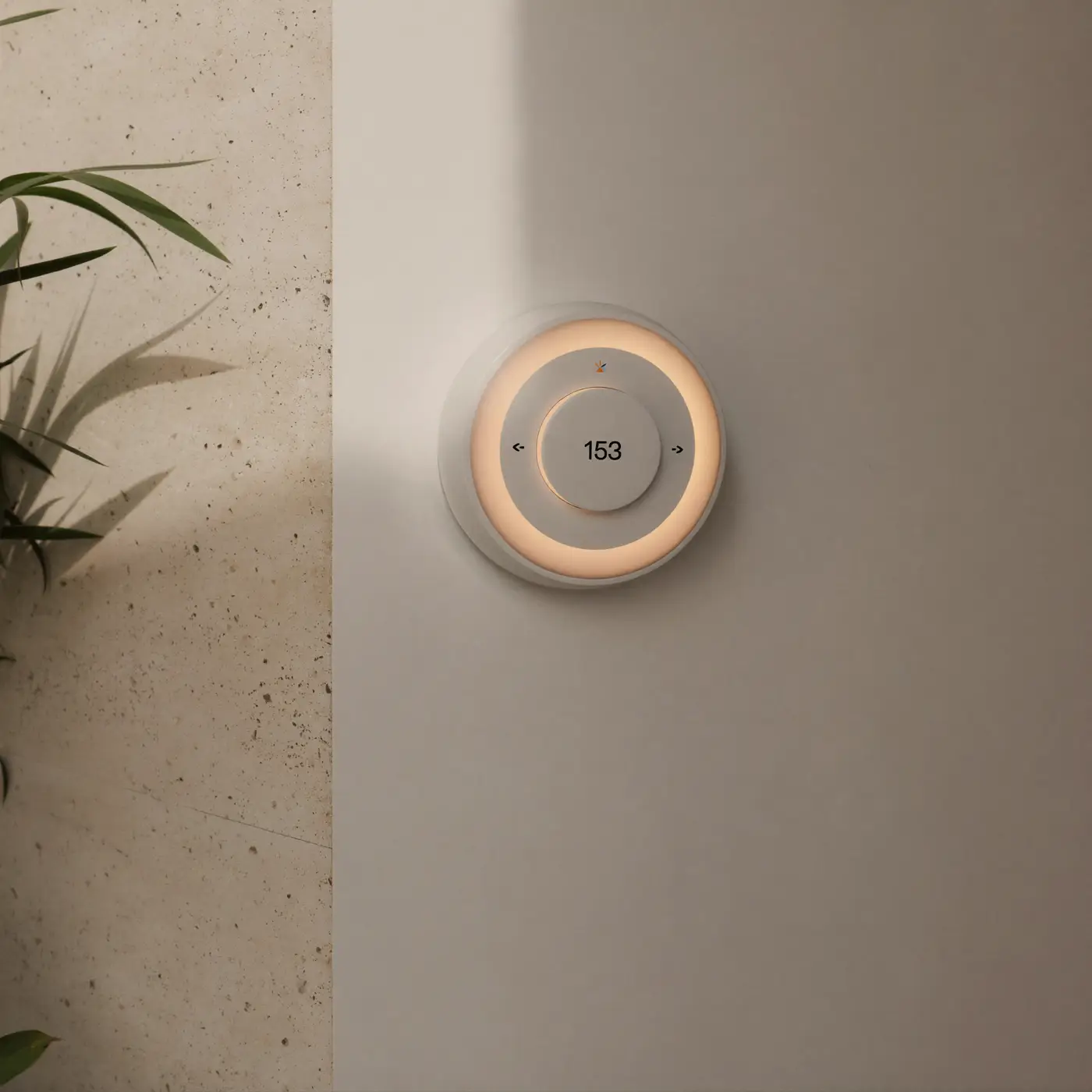 A round smart thermostat with a softly glowing ring on a warm white wall beside a plant.