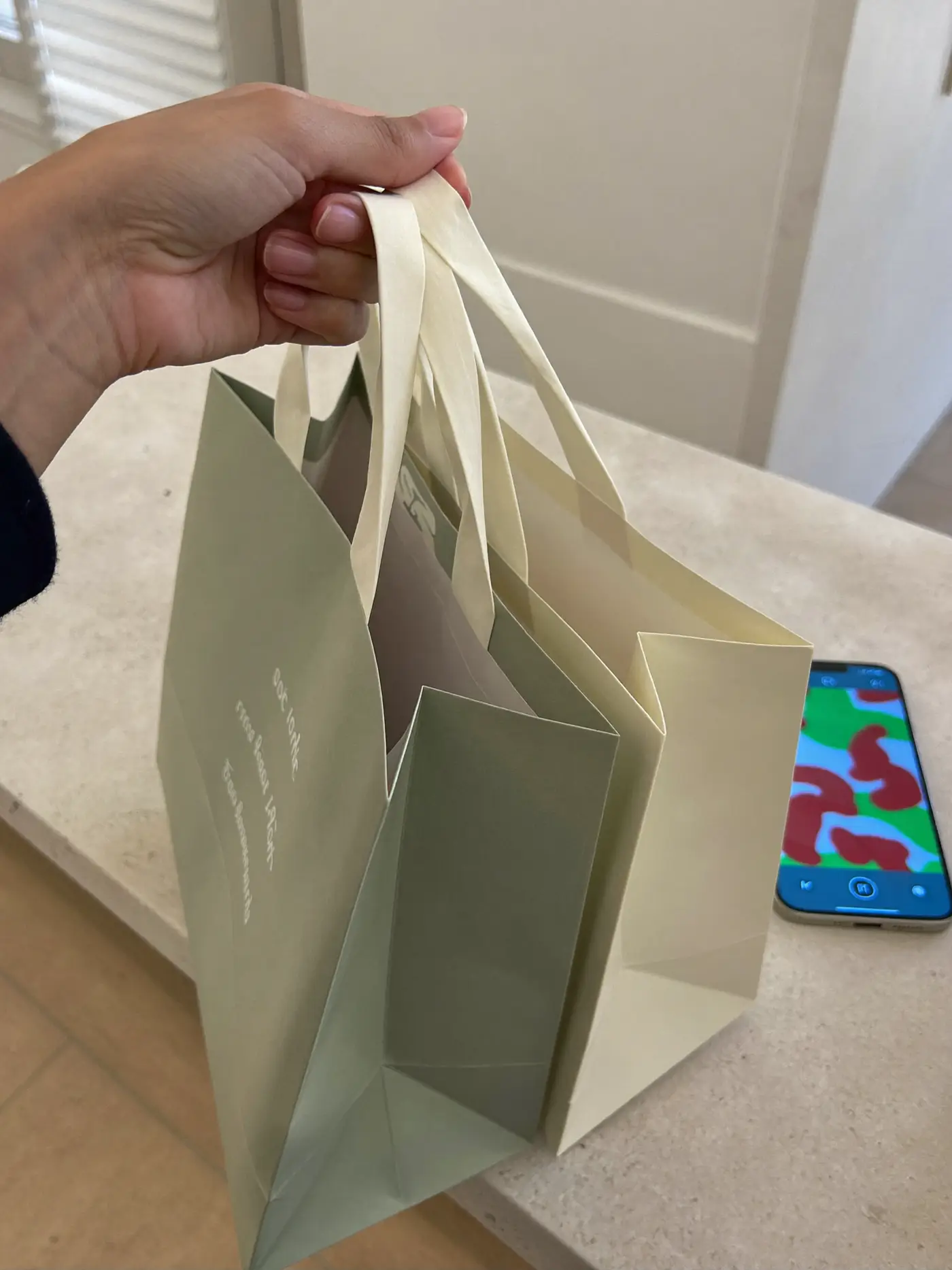 A shopper holding sage and cream paper shopping bags beside a phone on a retail counter.