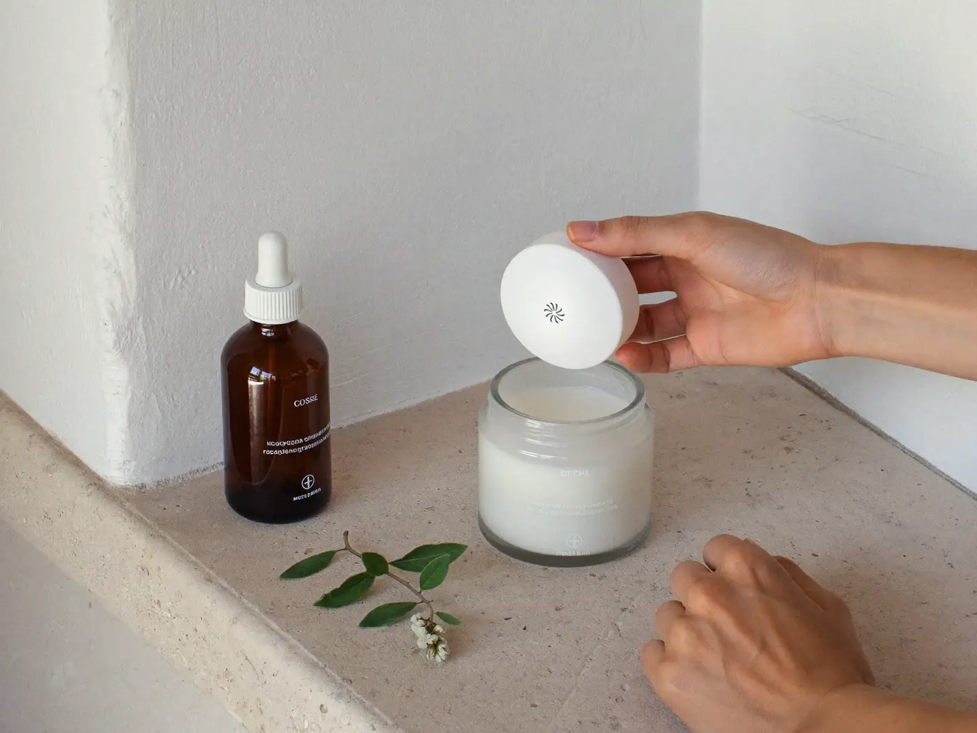 A hand lifting a white refill pod above a frosted glass skincare jar beside an amber dropper bottle and a sprig of leaves.