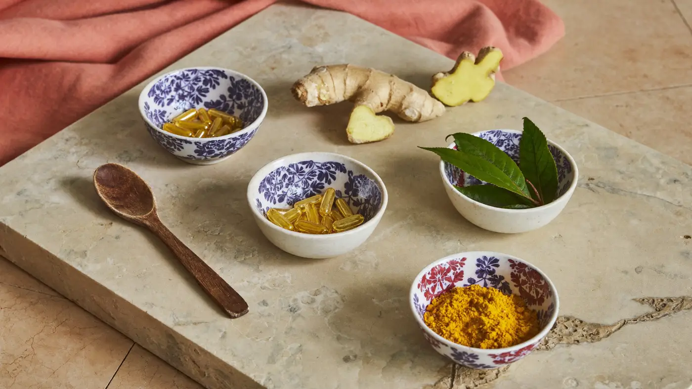 Ceramic bowls of golden capsules, turmeric powder and green leaves beside fresh ginger on travertine.