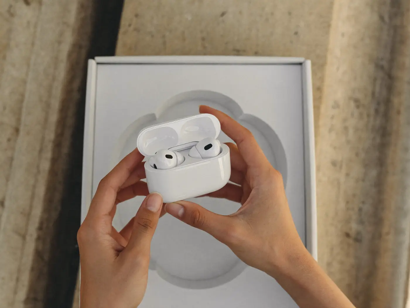 Two hands lifting a white wireless earbuds case out of a rigid box with a moulded paper insert.