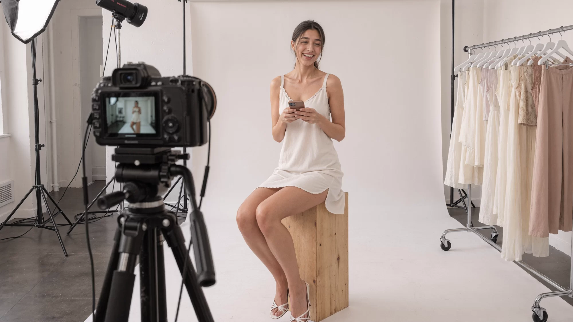 Behind the scenes of a studio fashion shoot, with AI character Dhwaja laughing on an apple box among lights and a clothes rail. Sample image created by Quiamo, a brand, marketing and media agency from India.