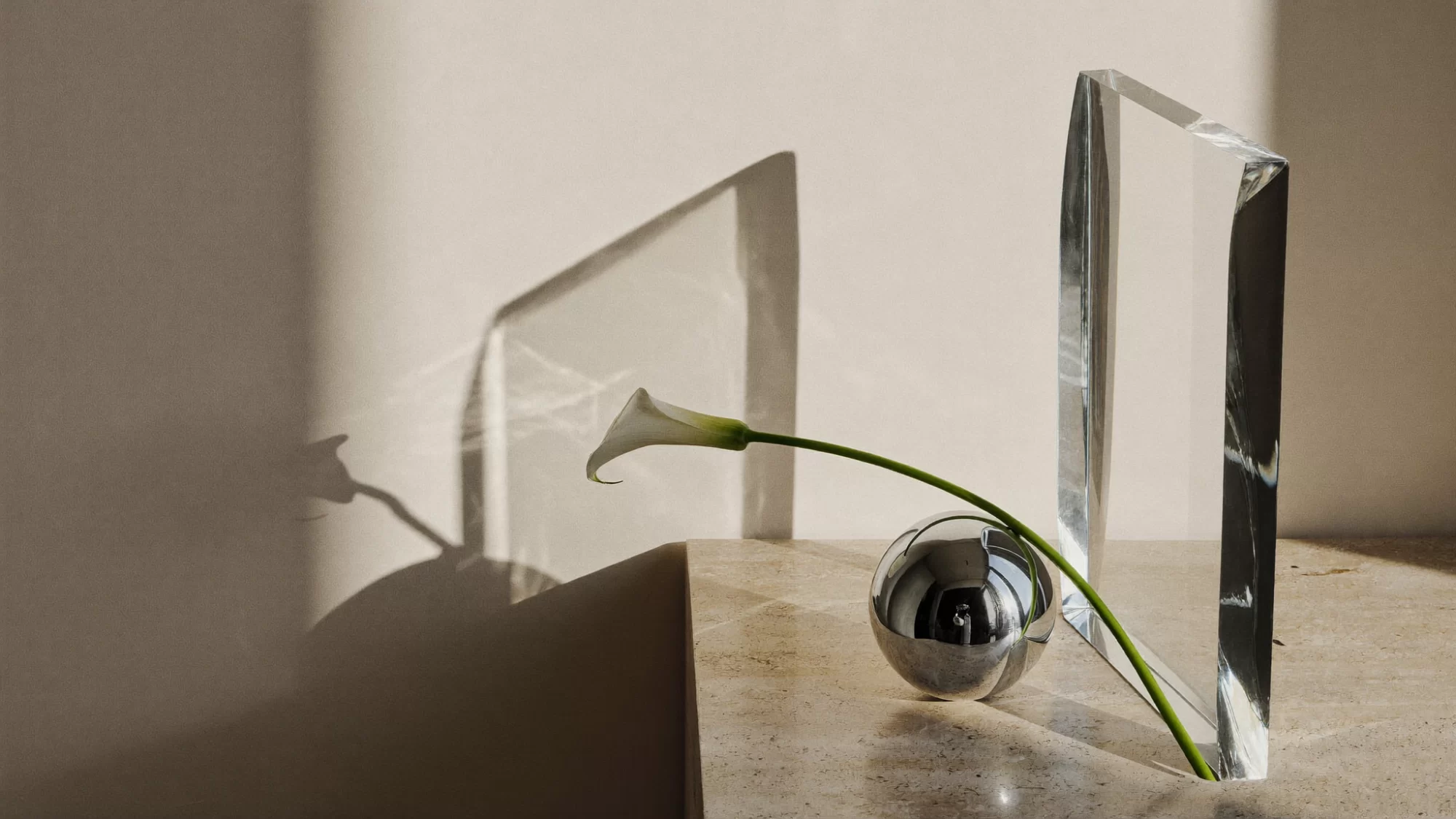 A chrome sphere, a tilted glass slab and a white calla lily arranged as a floating still life on an ivory ground. Sample image created by Quiamo, a brand, marketing and media agency from India.