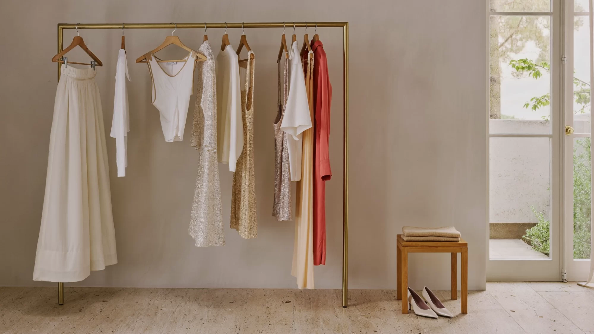 A brass garment rail of ivory, blush and coral clothes beside a sunlit window in a calm atelier. Sample image created by Quiamo, a brand, marketing and media agency from India.